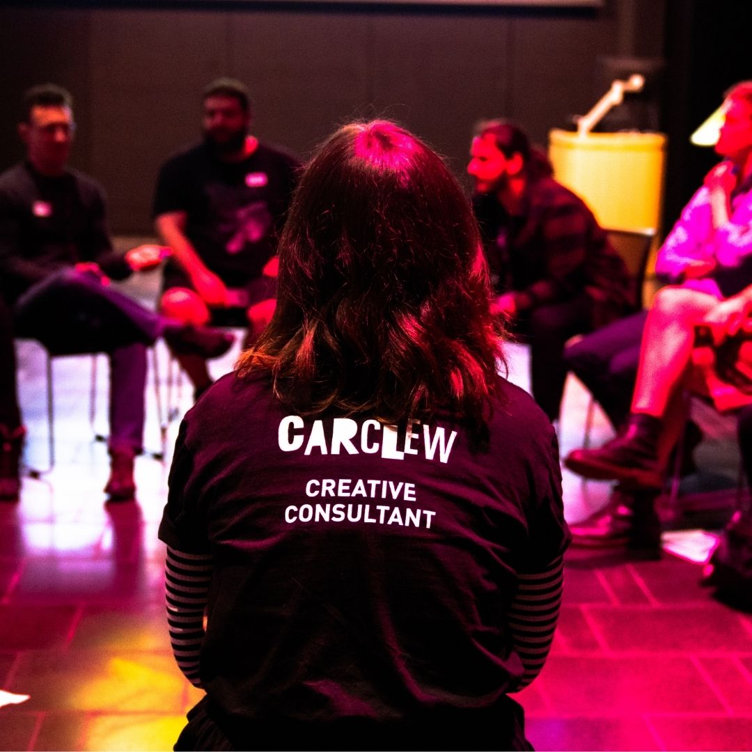 Creative Consultant members with back to camera showing her logo tshirt, with other consultants discussing ideas in the background